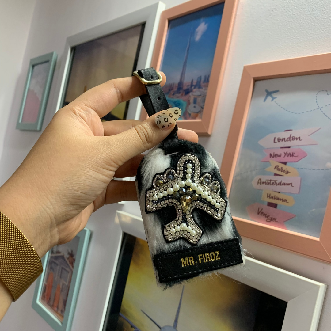 A hand holding a rectangular luggage tag with a polka dot pattern and a personalized name tag attached to it. The tag is black with silver accents, and it&