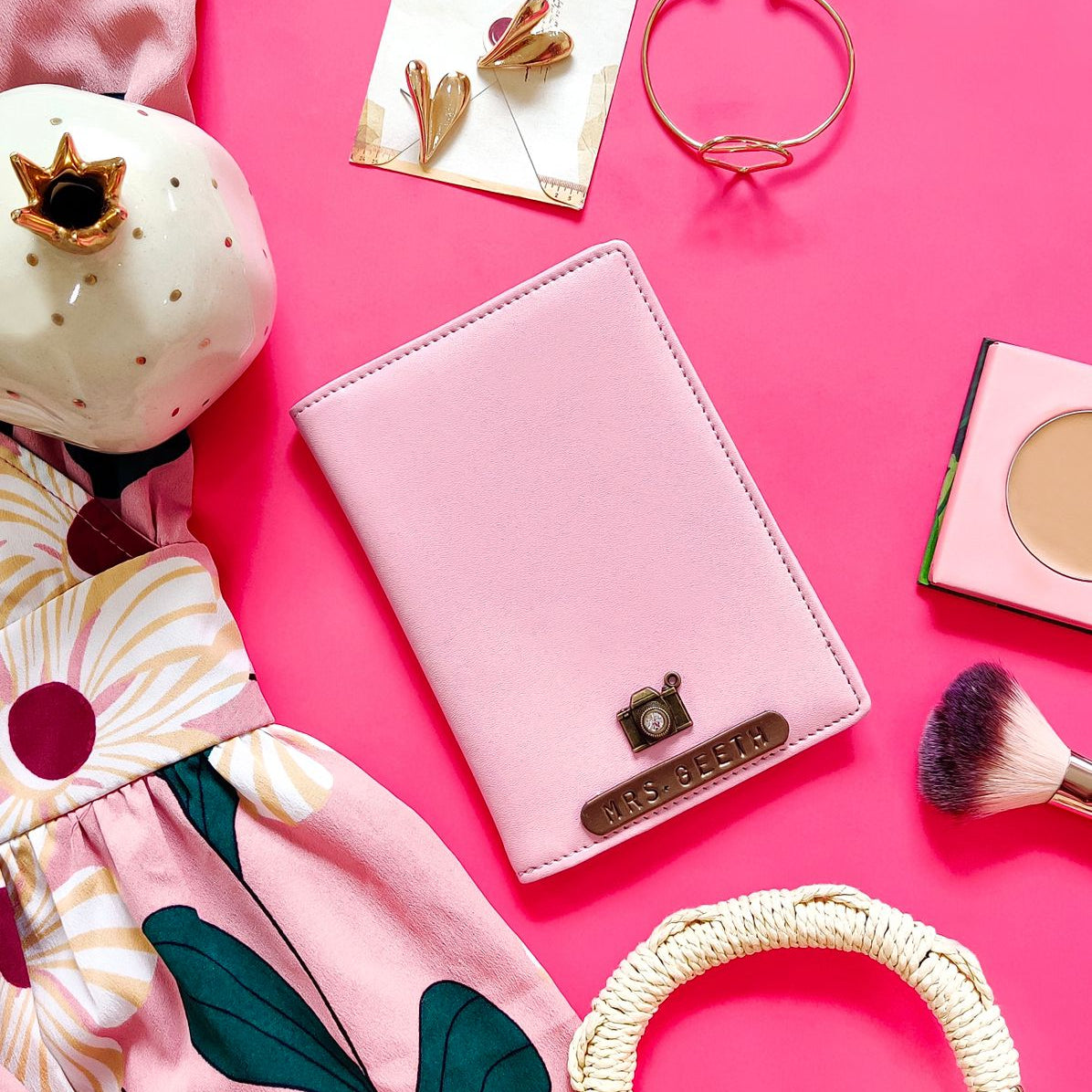 A passport cover in baby pink with a crocodile texture, alongside a name tag, charm, and other accessories on a pink background.