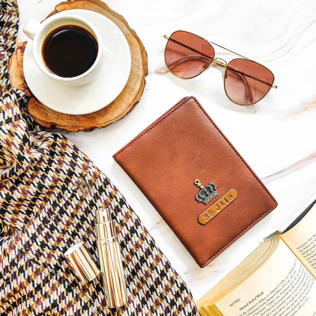 A brown passport cover with a name tag and charm, placed on a table alongside a cup of coffee, sunglasses, and a book.