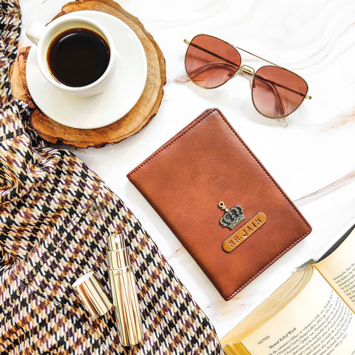 A brown passport cover with a name tag and charm, placed on a table alongside a cup of coffee, sunglasses, and a book.