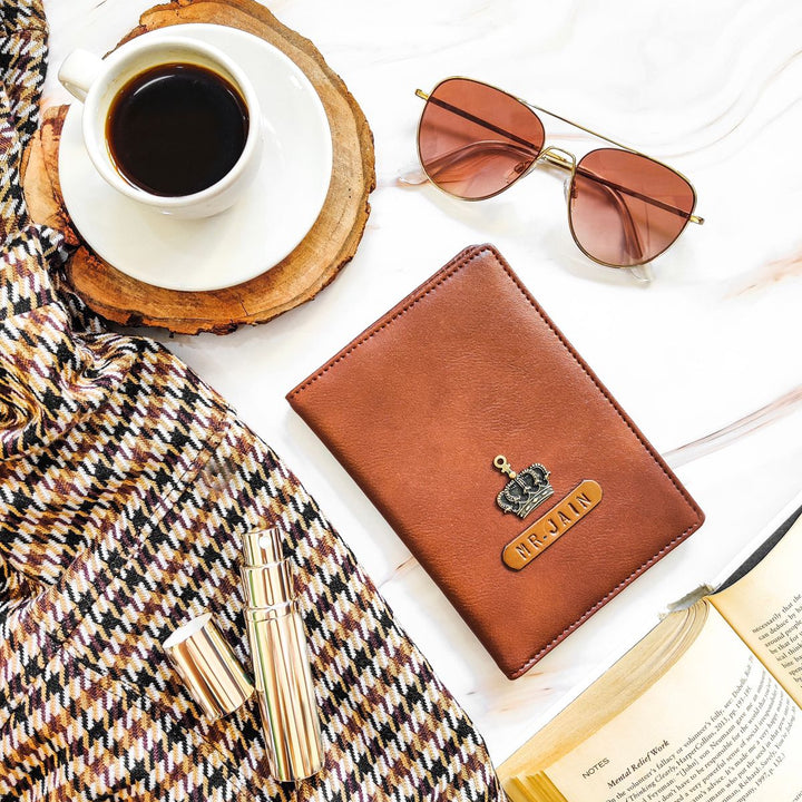A brown passport cover with a name tag and charm, placed on a table alongside a cup of coffee, sunglasses, and a book.