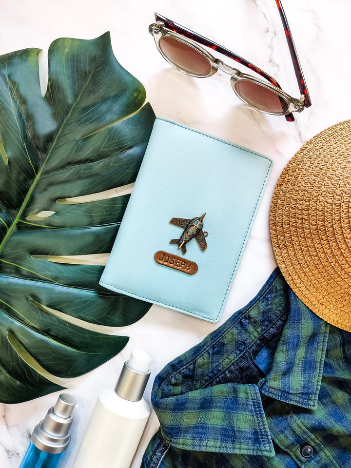 A light blue passport cover with a crocodile pattern, alongside a pair of sunglasses, a straw hat, and a name tag.