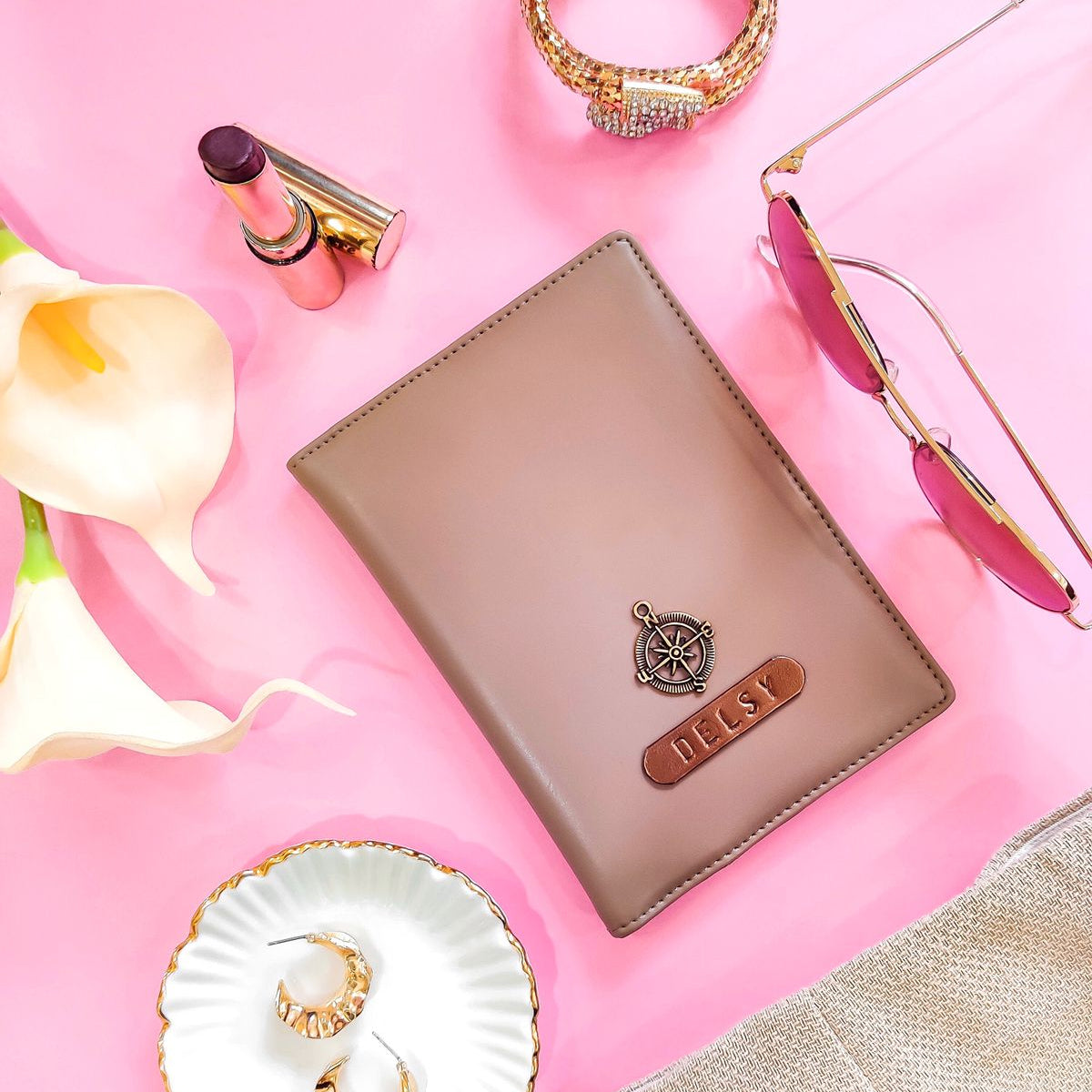 A brown passport cover with a crocodile pattern texture, displayed alongside a variety of accessories including a passport, sunglasses, and earrings on a pink background.