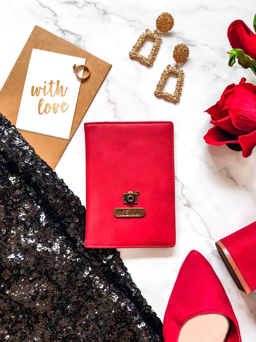 A red passport cover with a personalized name tag and charm, placed on a marble surface alongside a pair of earrings, a card, and a rose.