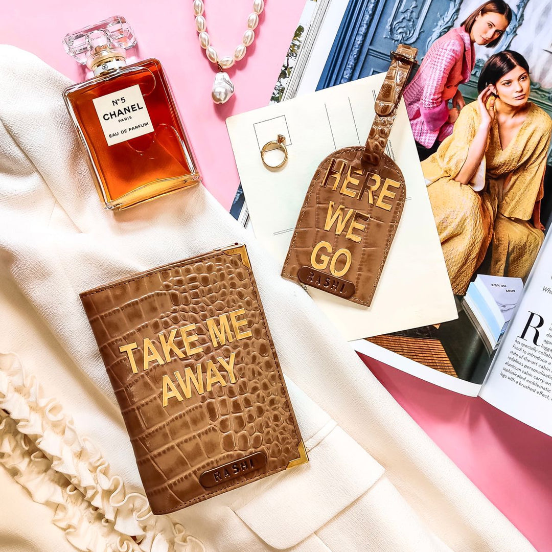 A set of genuine leather passport cover and luggage tag set with a name tag and charm, displayed alongside a perfume bottle and a fashion magazine. - TPC Gifts